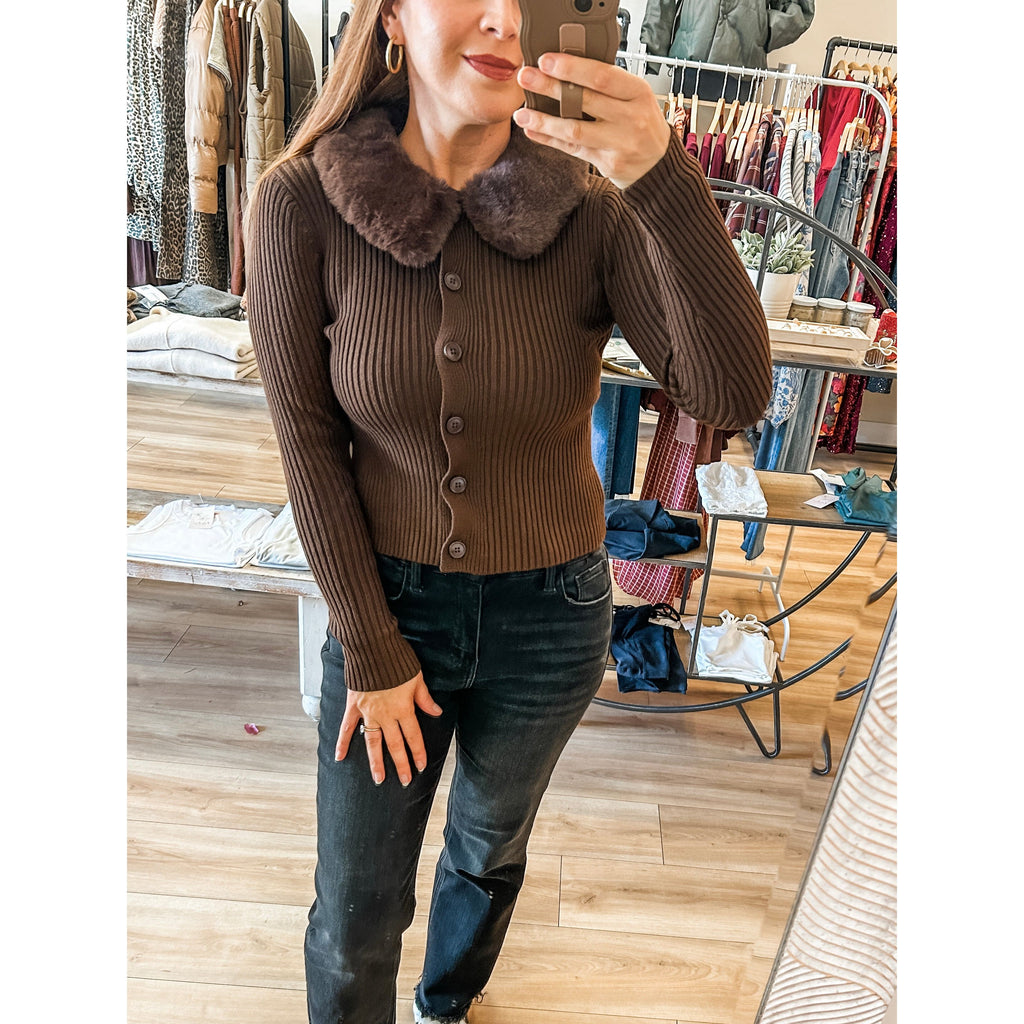 Person taking a mirror selfie wearing a brown sweater with a collar, dark jeans, and white sneakers in a clothing store.